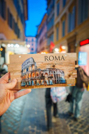 Téléchargez les photos : Main tenant une carte postale avec une représentation artistique du Colisée à Rome, Italie. Texture rustique en bois, ROMA et COLOSSEO affichés. Fond flou de la scène de rue avec des bâtiments européens traditionnels. - en image libre de droit