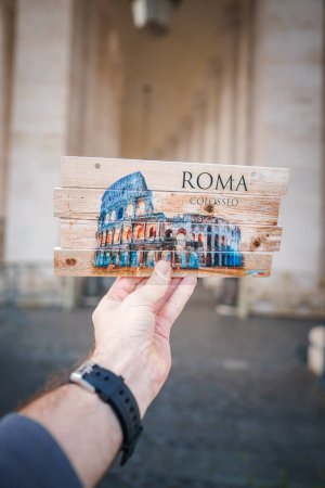 Foto de Colorido recuerdo que representa el icónico anfiteatro romano, el Coliseo, en madera. ROMA COLOSSEO impreso. Mano de la persona sosteniendo. Fondo exterior borroso. - Imagen libre de derechos