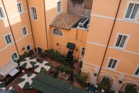 Innenhof eines Luxushotels in Rom mit warmen Pfirsichwänden, weißen Fenstern, Sonnenschirmen und Terrakottadächern. Gemütliche Sitzecke im Freien mit viel Grün und Brunnen schaffen eine intime urbane Oase.