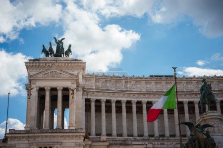 Altare della Patria, o Monumento Nacional a Víctor Manuel II en Roma, Italia. Gran edificio con columnas blancas, estatuas, esculturas de carros y bandera italiana. Día soleado destaca su fachada.
