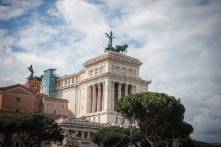 Landmark in Rome, Italy, featuring Altare della Patria a grand, white marble monument with statues of chariots symbolizing victory. Sky is partly cloudy in the background.