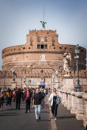 Téléchargez les photos : Les touristes se promènent le Ponte SantAngelo à Rome, en Italie, avec le Castel SantAngelo en toile de fond. La scène présente des statues d'ange, un ciel ensoleillé et une atmosphère détendue. - en image libre de droit