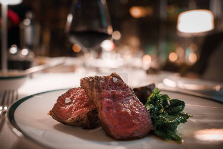 Close up view of succulent steak and green vegetables on an elegant plate. In a luxurious fine dining setting with soft lighting and a glass of red wine.