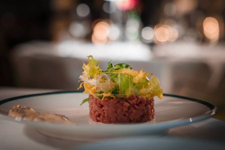 Elegant dish with steak tartare on white plate with blue rim in upscale restaurant setting. Vibrant colors, cozy ambiance, focus on luxury dining experience.