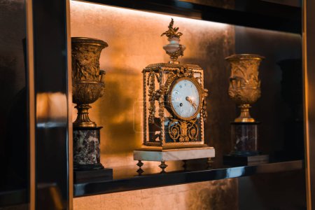 Opulente antike Uhr, Urnen mit klassischem Design, luxuriöses Dekor im Hotel in Rom, aufwändige Metallverarbeitung, weißes Zifferblatt, elegante Details, warme Beleuchtung.