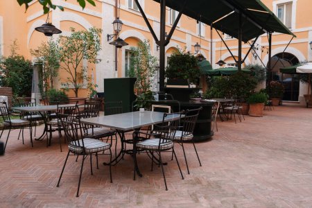 Luxuriöser Sitzbereich im Innenhof des Hotels in Rom. Terrakottafliesen, pfirsichfarbene Wände, eiserne Möbel, grüne Sonnenschirme, Pflanzen und Laternen schaffen ein ruhiges Ambiente.