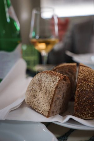 Frisch gebackenes Brot mit rustikalem Aussehen, präsentiert in einem Korb mit weißem Tuch. Im Hintergrund ein Glas Weißwein. Wahrscheinlich in einem Luxushotel in Rom.