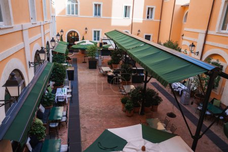 Luxushotelhof in Rom europäische Architektur, warme Wände, gemauertes Pflaster, Topfpflanzen. Tische zum Essen unter grünen Sonnenschirmen. Heitere, ruhige Atmosphäre.