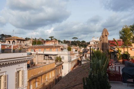 Malerischer Blick auf die Dachterrasse vom Luxushotel in Rom. Gartenmöbel, grüne Pflanzen, sonniges Stadtbild mit warmgetönten Gebäuden und italienischen Kiefern. Gelassene Atmosphäre.