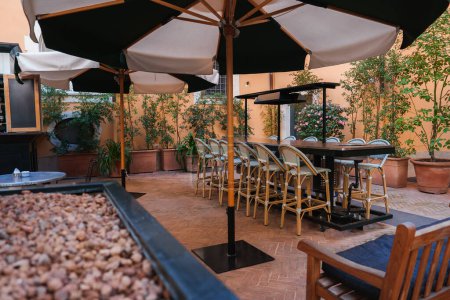 Luxury hotel outdoor seating area in Rome. Warm ambiance with terracotta tiles, cream umbrellas, rattan chairs, lush plants. Elegant and tranquil setting.