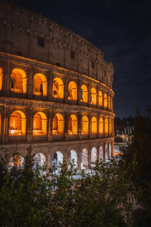 Ancient Colosseum in Rome illuminated by warm golden lights at night, showcasing iconic arches and outer walls. Dark sky suggests late evening. Iconic landmark in Rome, Italy.