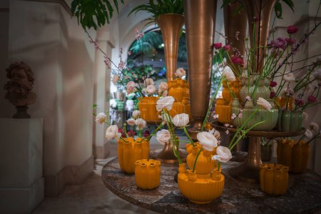 Arrangement floral luxueux dans un restaurant haut de gamme. Roses en blanc, rose et fuchsia. Vases aux teintes jaunes à vertes sur table en mosaïque. Décor élégant avec colonnes et buste sculpture.