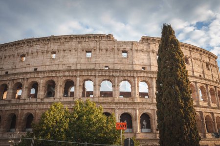 Antikes Kolosseum in Rom, Italien. Tagesaufnahme eines gut erhaltenen Amphitheaters unter teilweise bewölktem Himmel. Historische Architektur mit modernem Taxistand-Schild im Vordergrund.