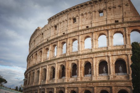 Vue rapprochée du célèbre Colisée elliptique de Rome, en Italie. Les arcs de pierre, les statues et l'importance historique sont capturés sous un ciel chaud et nuageux. Les travaux de préservation en cours sont visibles.