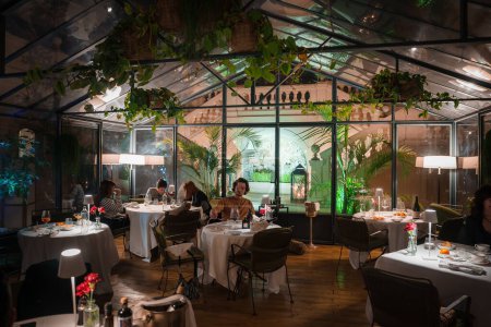 Luxuriöses Restaurant-Interieur mit Glaswänden und -decken, üppigem Grün, eleganten Speiseeinrichtungen und gemütlicher Beleuchtung. Hochwertiges kulinarisches Erlebnis in anspruchsvollem und heiterem Ambiente.