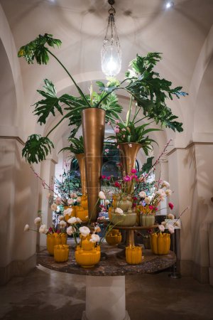 Elegante arreglo floral en lujoso restaurante con jarrón de oro metálico, exuberante follaje verde, flores rosadas, orquídeas blancas y opulentos detalles de decoración..