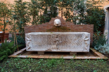 Fuente de piedra con diseño clásico y cabeza de león en un jardín sereno en un hotel de lujo en Roma. Características esculturas mitológicas en relieve y exuberante vegetación, evocando el encanto histórico.