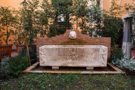 Ruhiger Innenhof in einem Luxushotel in Rom. Ein reich verzierter Brunnen aus Stein mit Löwenkopf ergießt Wasser in ein Becken, das von üppigem Grün und Pfirsichmauern umgeben ist. Elegantes und ruhiges Ambiente.