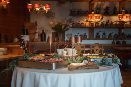 Salle à manger rustique à l'ambiance chaleureuse avec table à buffet avec viandes, fromages et ustensiles de cuisine vintage dans un décor traditionnel de style alpin ou champêtre.