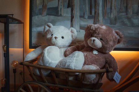 Cozy teddy bears in vintage carriage in a warm, inviting interior. One white, one brown with abstract painting background. Display exudes comfort and nostalgia ambiance.