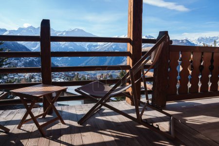Holzbalkon mit Bergblick und dekorativem Geländer. Tisch und Stuhl zum Entspannen oder Essen. Schneebedeckte Berge, unten eine kleine Stadt. Sonniges Tagambiente. Friedliches Umfeld.