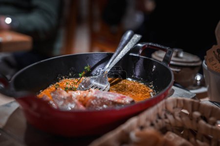 Kochszene mit einer Nahaufnahme einer roten Pfanne auf Herdplatte mit angebratenem oder gebratenem Essen. Frische Kräuter darauf mit einer Zange drinnen. Küchenumgebung im Hintergrund.