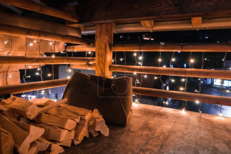 Indoor setting with rustic, cozy ambiance. Wooden box filled with firewood in foreground, warm lights above. Could be mountain lodge or countryside retreat.