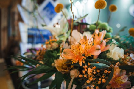Colorido arreglo floral con vibrantes azucenas de color naranja, pequeñas flores de color naranja, botón amarillo como flores, y variada vegetación en un ambiente interior borroso.