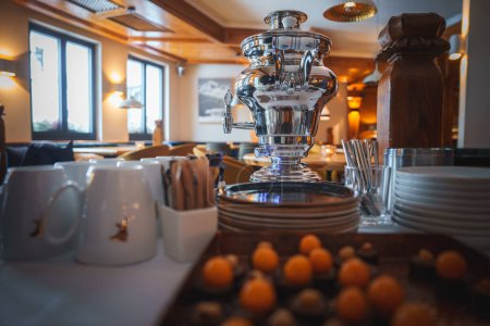 Elegant buffet setup with shiny silver samovar, white plates, wooden pepper mill, ceramic mugs, wooden utensils, kumquats, warm interior atmosphere. Ideal for meal service.