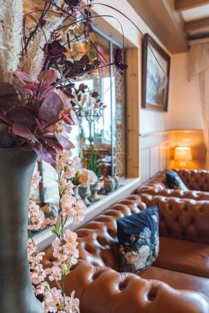 Elegantes Wohnzimmer mit großer Vase mit künstlichen Blumen auf der Fensterbank, Chesterfield-Ledersofa, gerahmten Kunstwerken und gemütlicher Beleuchtung. Komfort und Raffinesse.