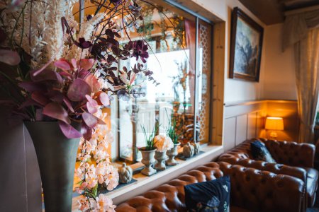 Gemütliches Eckzimmer-Dekor mit leuchtenden rosa, lila und weißen Blumen in einer großen Vase, gemustertem Vorhang, Chesterfield Sofa und eklektischen Dekorationselementen. Reiches, luxuriöses Gefühl.