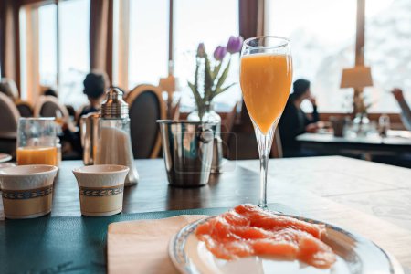 Dining-Szene in einem modernen Interieur, mit einem Glas orangefarbenem Getränk, Schüsseln mit blauem Goldrand, Eiskübel, geräuchertem Lachs und Gästen, die Mahlzeiten genießen.