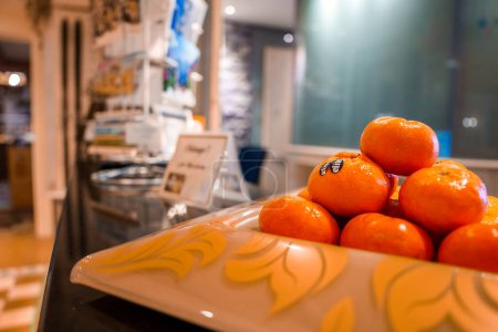 Close up view of bright orange tangerines on a yellow patterned plate, set on a black countertop in a cozy interior space with blurred background details.