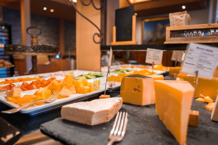 Assortment of cheeses on buffet table, Swiss style wedges with holes, soft rindless varieties, cheddar chunks on skewers, sliced cucumbers, signs, warm ambiance, wood tones.