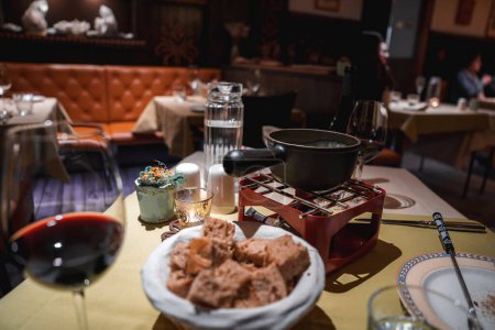 Dinner-Szene im einladenden Restaurant Tisch für Mahlzeit w oder Wein, Kerze, Pflanztopf, Fondue-Topf auf rotem Ständer, Brotkorb gedeckt. Intimes, gemütliches Ambiente.