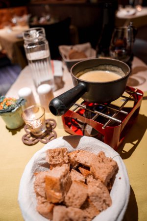 Cadre confortable avec panier à fondue de cubes de pain, variété à grains entiers, casserole à fondue sur support rouge avec mélange crémeux, verre à vin, salières, intérieur chaud, atmosphère intime.