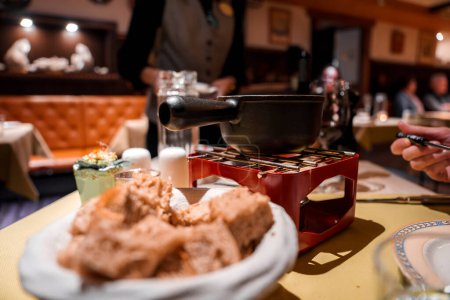 Foto de Escena de comedor con cubos de pan de configuración fondue tradicional en el plato, olla fondue negro en soporte rojo con quemador, comedor interactivo. Ambiente acogedor restaurante, sin ubicación identificada. - Imagen libre de derechos