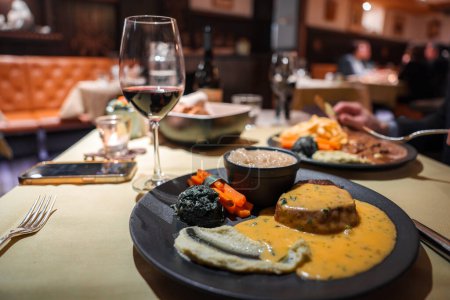 Dinner-Szene mit Gourmet-Steak und Gemüse auf Teller, Glas Rotwein und gemütlichem Ambiente. Smartphone und Gabel auf dem Tisch. Andere Gäste im Hintergrund.