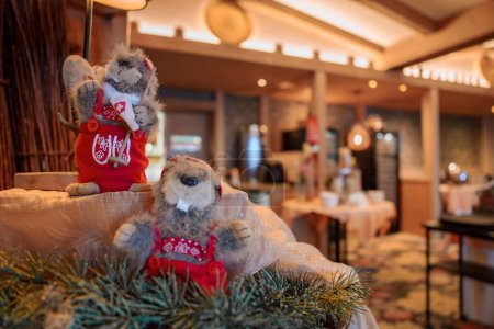 Two festive plush toy squirrels in warm, cozy indoor setting one in red dress with white floral patterns holding a mini ski, other in focus with baguette, possibly a restaurant or cafe ambiance.