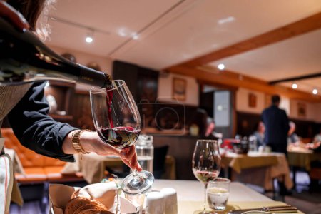 Person, die in einem eleganten Rahmen Rotwein in ein Weinglas gießt. Hintergrund zeigt gehobenen Essbereich mit gemütlichem Ambiente. Ideal für Weinauswahlunternehmen.