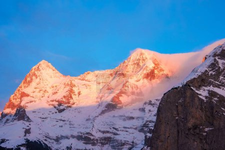 Photo for Breathtaking view of snow covered peaks at Murren ski resort, Switzerland at sunrise or sunset. Sun bathes mountains in pinkish orange light, contrasting with blue sky. Majestic alpine scenery. - Royalty Free Image