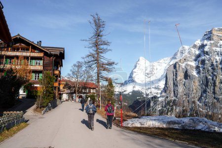 Photo for Alpine scene at Murren ski resort, Switzerland. People stroll on road past Swiss chalet as snowy mountains tower under clear sky, trees scattered around. - Royalty Free Image