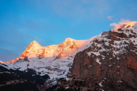 Atemberaubender Blick auf schneebedeckte Gipfel, Skigebiet Murren, Schweiz. Sonnenuntergang oder Sonnenaufgang beleuchten schroffe Gipfel, Schnee, Felsen, Wolken, ruhigen Himmel, bewaldete Hänge. Heitere natürliche Schönheit.