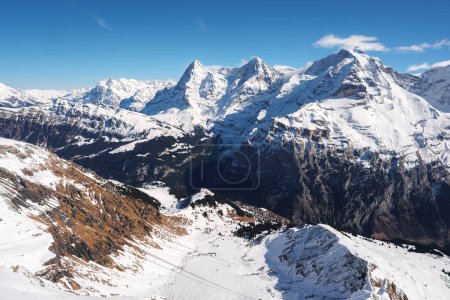 Breathtaking snowy peaks and rugged terrain of Murren ski resort in Switzerland. Snow covered slopes, ski lift cables, deep valley, forest patches, majestic Swiss Alps view under a clear blue sky.