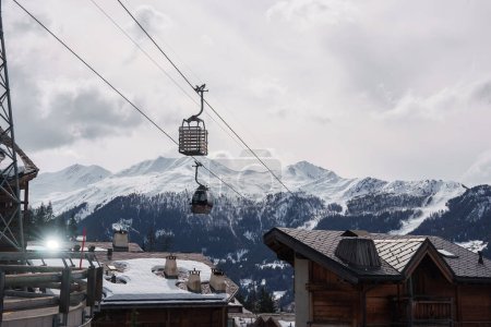 Winterlandschaft in einem Skigebiet mit Holzchalet-Gebäuden, Skilift, schneebedeckten Bergen und Skipisten. Ruhige und kühle Atmosphäre. Befindet sich in den europäischen Alpen oder einer ähnlichen Region.