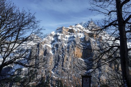 Majestätische Berggipfel in den Schweizer Alpen, wahrscheinlich in der Nähe von Murren Skigebiet. Schneebedecktes, zerklüftetes Gelände mit kahlen Bäumen im Vordergrund. Sonniger Tag mit einer Uhr, die eine menschliche Note verleiht.