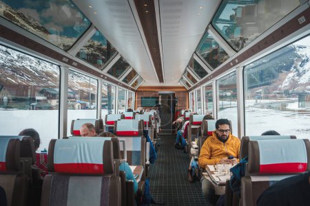 Photo for Luxurious train car interior on Glacier Express in Switzerland. Large windows offer unobstructed views of snow capped mountains, village. Comfortable seating, relaxed atmosphere, engaged passengers. - Royalty Free Image