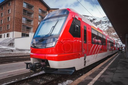 Téléchargez les photos : Train rouge moderne au quai, probablement partie de Glacier Express en Suisse. Design élégant avec grande fenêtre avant pour une vue large. Fond montagneux enneigé près de Zermatt. - en image libre de droit