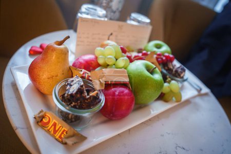 Gemütliche Obstplatte mit reifen Birnen, Trauben, Äpfeln, Johannisbeeren, Schokoriegeln und Müslimix auf Marmortisch im Luxushotel in Zermatt, Schweizer Skiort.