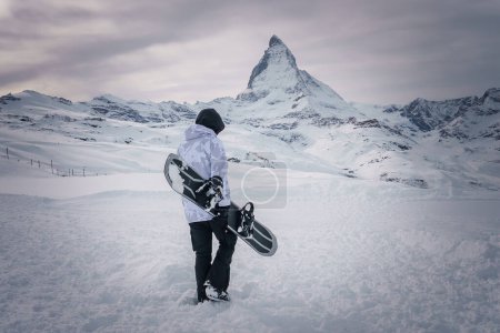 Snowboarder at Zermatt ski resort, facing Matterhorn mountain. Dressed in winter gear and holding a snowboard with bindings. Serene and contemplative scene with anticipation for adventure.